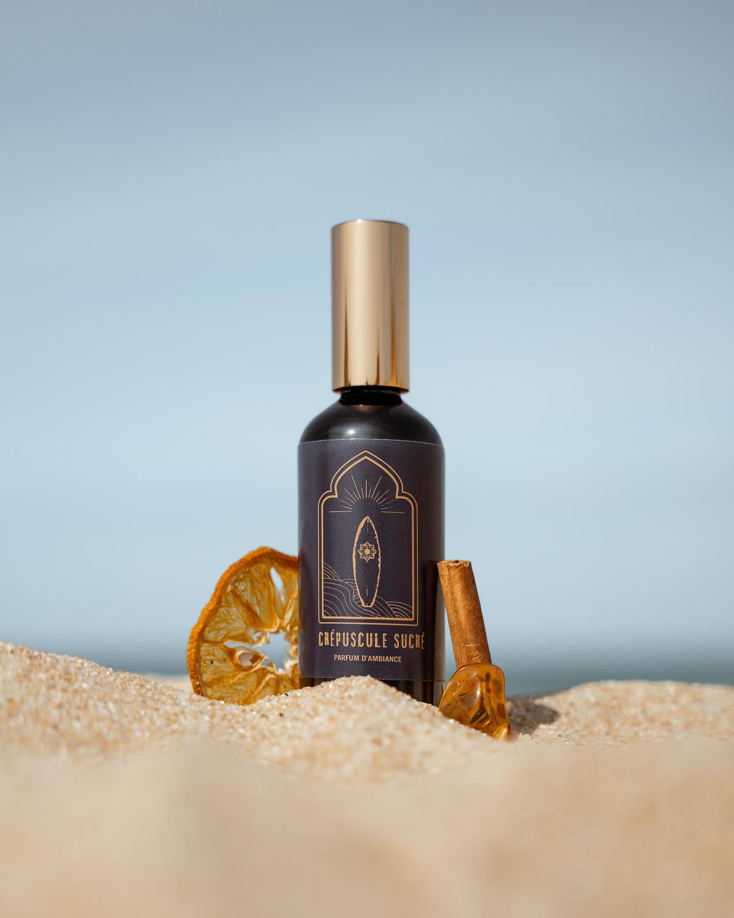 Parfum d'ambiance Mawja crépuscule sucré, flacon verre de face sur la plage dans le sable avec orange séché et bâton de cannelle